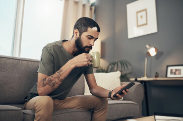 Connect to all things current with the ease of technology. Shot of a young man having coffee and using a smartphone on the sofa at home.