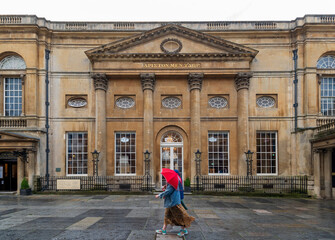 City of Bath, UK. People with red umbrella walking in front of restored in Victorian times ancient Roman Baths.  The Pump Room. Translation from old Greek: Water is Best.