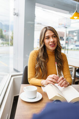 young latin girl with blonde hair makeup smiling, with an open book, pencil and a cup of hot drink...