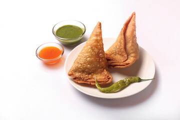 Two samosas kept on a white dish with chilly. Red & green chutney ket in a bowl.