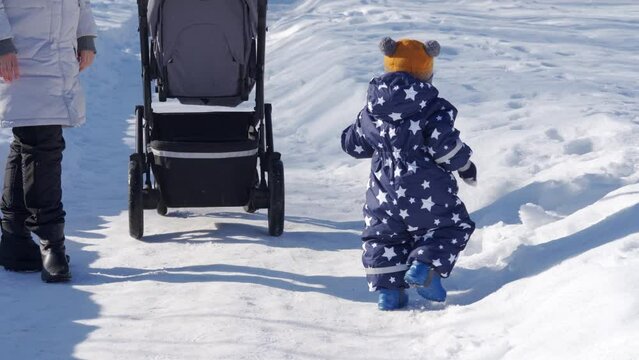 Kid takes the first steps in the snow walking with mom. 1 year old baby boy learning to walk with mom in the park in winter. High quality 4k footage