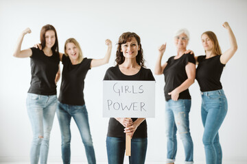 Four women dressed in jeans and black T-shirts raise their hands in a gesture of strength and fifth woman standing with tab with the inscription Girls power
