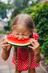 Funny little girl in red dress eating watermelon in the garden.