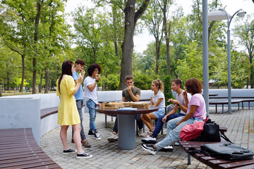 Group of young people gathering around table in park and enjoying pizza, bags and backpacks lying on bench beside. Arc shot zoom out students having lunch together after classes. Concept of food