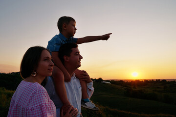 Family standing on hill in countryside at sunset. Arc shot little boy sitting on shoulders of father and pointing at something in the distance, mother standing beside. Concept of nature