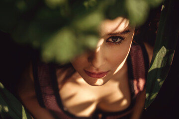 A close-up portrait of a woman in a low key in a hard light, one eye is covered with the leaves of a geranium plant, on the woman's face there are sun glare and shadows from plants
