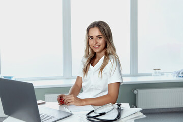 Smiling female doctor is sitting at desk - obrazy, fototapety, plakaty