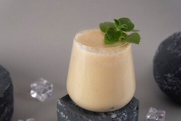 Glass of traditional Indian lassi drink with mint leaf, mango and ice on a grey background.