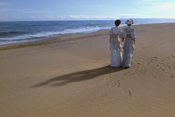 Two women in victorian fashion strolling along a beach - obrazy, fototapety, plakaty