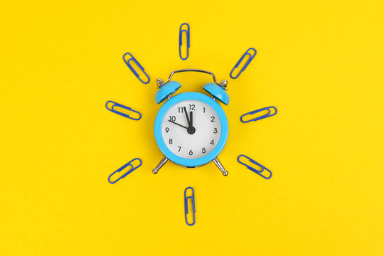 On the yellow surface lies a blue alarm clock and paper clips - depict an alarm clock.