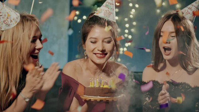 Portrait of attractive asian girl blowing out fire candles on birthday cake under falling glowing confetti. Girlfriends smiling, laughing, rejoicing, congratulating and claping birthday girl.