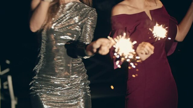 Asian and caucasian girls dancing together, waving lighted sparklers. Happy girlfriends relaxing and enjoying celebration in aning or night. Cheerful women having fun alone at festive party.