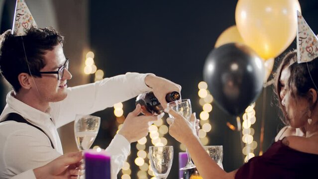 Guys celebrating Party Party or Party, smiling, laughing, talking and communicating. Millennial friends at party sitting at table, opening bottle of champagne and pouring sparkling wine into glasses.