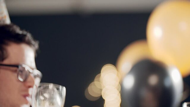 Portraits of young multiracial girls sitting at festive table and celebrating holiday. Women congratulating friends, smiling, laughing, blowing whistle, clinking glasse of champagne and drinking wine.