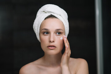 Closeup head shot pleasant beautiful woman applying moisturizing creme on face after shower. Young pretty lady wrapped in towel smoothing perfecting skin, daily morning routine concept.