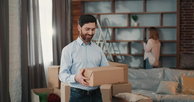 Portrait of happy man receiving home delivery in card box from postal worker, standing in spacious room after relocation 