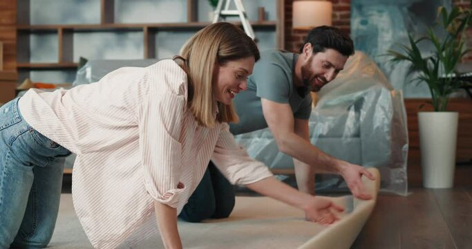 Happy caucasian couple rolling out carpet on floor, while making repair in apartment 