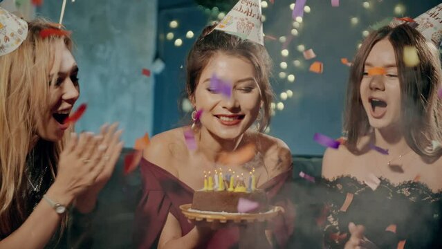 Portrait of attractive asian girl blowing out fire candles on birthday cake under falling glowing confetti. Girlfriends smiling, laughing, rejoicing, congratulating and claping birthday girl.