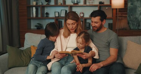 Loving mother and father reading book with two kids and sitting at cozy living room