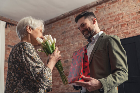 senior grey-haired caucasian woman getting a flower bouquet and a gift from her son on Mother's Day. High quality photo