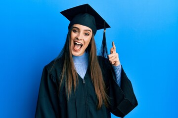 Beautiful brunette young woman wearing graduation cap and ceremony robe pointing finger up with successful idea. exited and happy. number one.