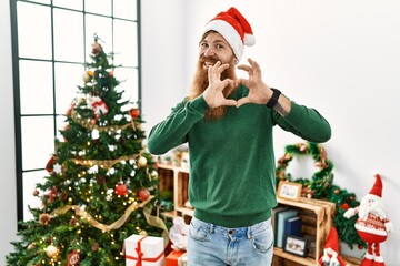 Redhead man with long beard wearing christmas hat by christmas tree smiling in love doing heart symbol shape with hands. romantic concept.