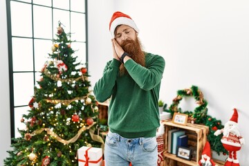 Redhead man with long beard wearing christmas hat by christmas tree sleeping tired dreaming and posing with hands together while smiling with closed eyes.