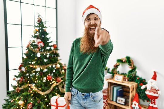 Redhead man with long beard wearing christmas hat by christmas tree pointing displeased and frustrated to the camera, angry and furious with you