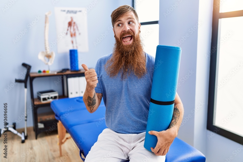 Sticker Redhead man with long beard holding yoga mat at rehabilitation clinic pointing thumb up to the side smiling happy with open mouth