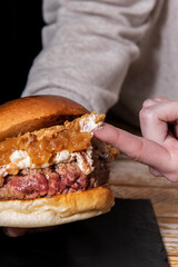 Holding in two hands an amazing beef burger with a lot of cream cheese and caramelized onions dipping the index finger in the cream cheese