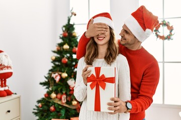 Young hispanic couple wearing christmas hat. Man surprising his girlfriend with hands on eyes and gift at home.