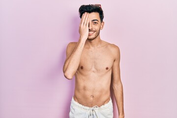 Fototapeta premium Young hispanic man wearing swimwear shirtless covering one eye with hand, confident smile on face and surprise emotion.