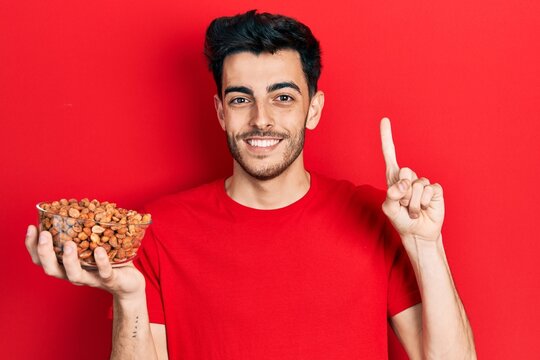 Young hispanic man holding peanuts smiling with an idea or question pointing finger with happy face, number one