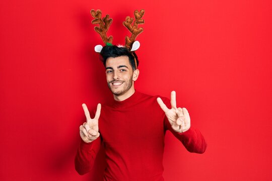 Young hispanic man wearing cute christmas reindeer horns smiling looking to the camera showing fingers doing victory sign. number two.