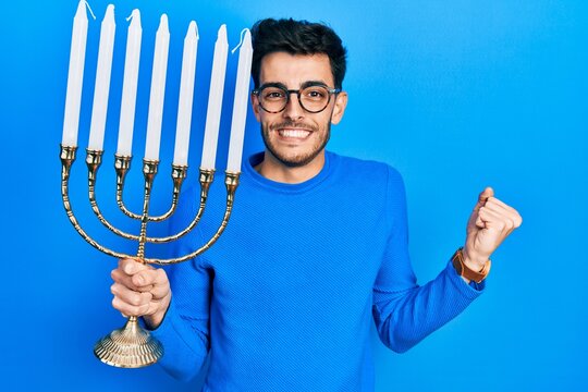 Young hispanic man holding menorah hanukkah jewish candle screaming proud, celebrating victory and success very excited with raised arm