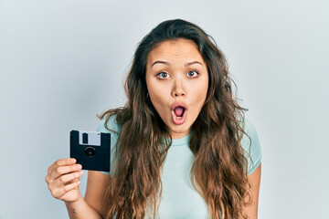 Young hispanic girl holding floppy disk scared and amazed with open mouth for surprise, disbelief...