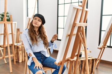 Young hispanic artist woman painting on canvas at art studio with hand on chin thinking about question, pensive expression. smiling with thoughtful face. doubt concept.