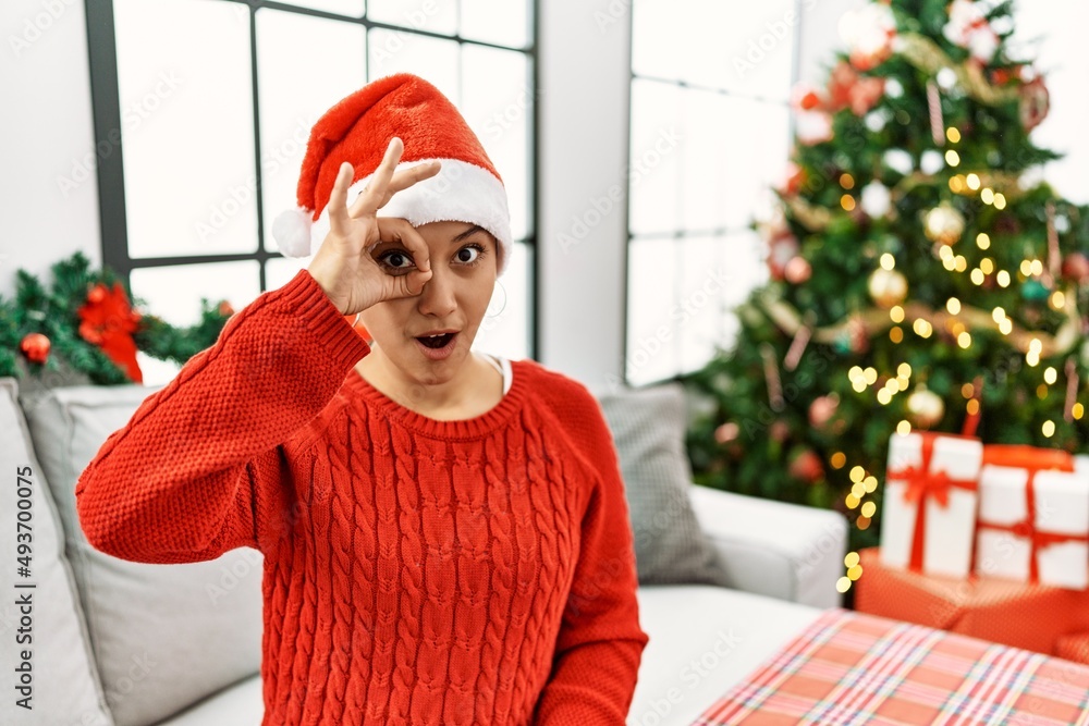 Wall mural Young hispanic woman with short hair wearing christmas hat sitting on the sofa doing ok gesture shocked with surprised face, eye looking through fingers. unbelieving expression.