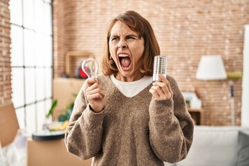 Young beautiful woman holding led lightbulb and incandescent bulb angry and mad screaming...