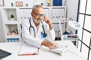 Fototapeta na wymiar Senior grey-haired man wearing doctor uniform talking on the telephone at clinic