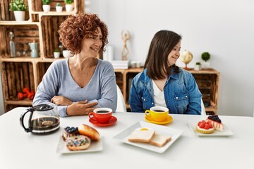 Family of mother and down syndrome daughter sitting at home eating breakfast looking away to side with smile on face, natural expression. laughing confident.