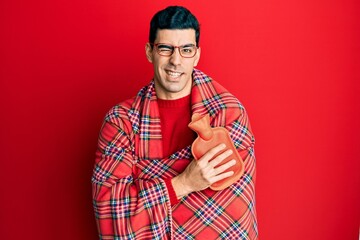 Handsome hispanic man wrapped in a winter blanket holding hot water bag winking looking at the...