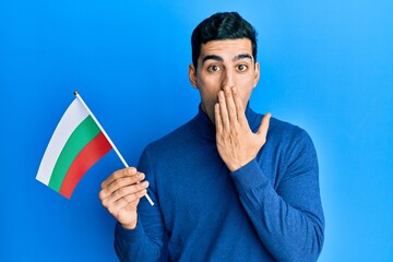 Handsome hispanic man holding bulgarian flag covering mouth with hand, shocked and afraid for...