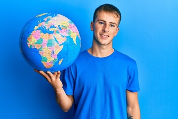 Young caucasian man holding world ball looking positive and happy standing and smiling with a confident smile showing teeth