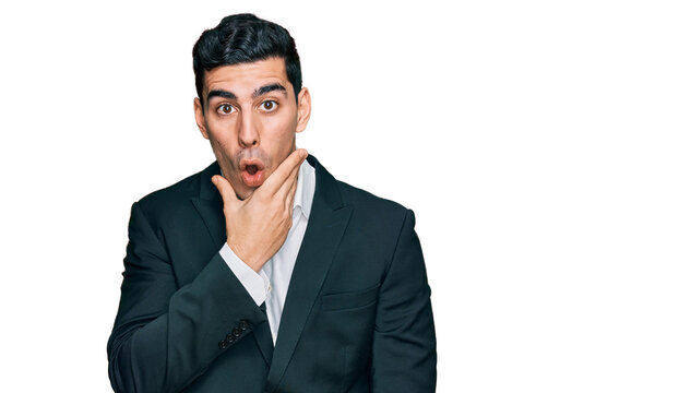 Handsome hispanic man wearing business clothes looking fascinated with disbelief, surprise and amazed expression with hands on chin