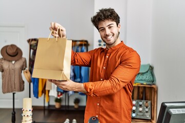 Young hispanic shopkeeper man smiling happy holding shopping bag at clothing store.