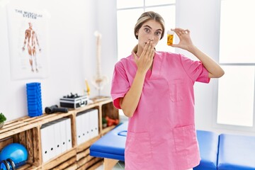Young physiotherapist woman working at pain recovery clinic holding pills covering mouth with hand, shocked and afraid for mistake. surprised expression