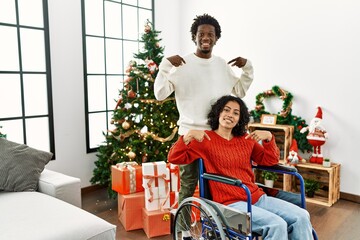 Young interracial couple with woman sitting on wheelchair by christmas tree looking confident with smile on face, pointing oneself with fingers proud and happy.