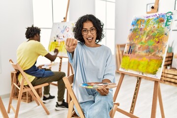 Young hispanic woman at art studio pointing to you and the camera with fingers, smiling positive and cheerful