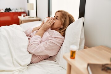 Middle age blonde woman smiling happy lying on the bed at home.